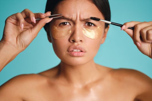 Close up Asian girl with patches under eyes seriously looking in camera with tweezers and brow brush